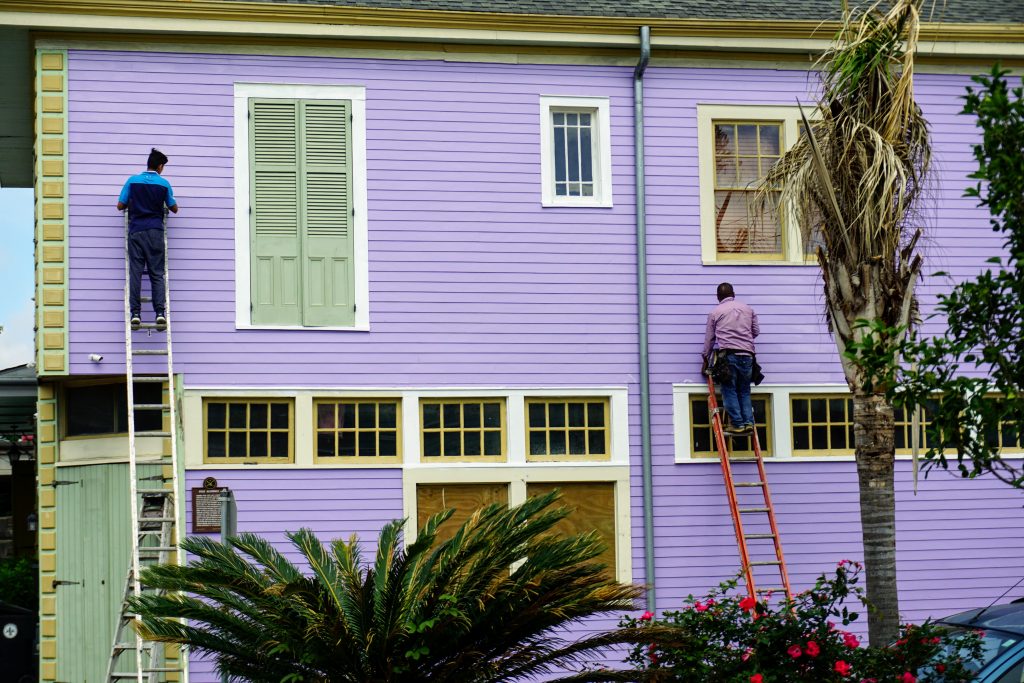 The 5 Biggest Home Renovation Costs to Sell a Home 3 Two men on ladders painting the exterior of a purple building — one of the priciest home renovation costs there is.